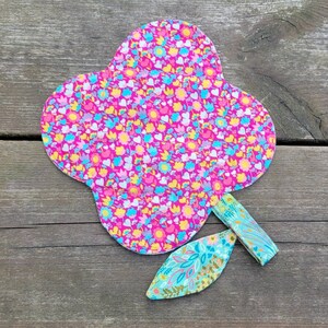 Pu&ograve; includere: Un fiore di stoffa rosa con un motivo floreale e uno stelo di stoffa verde. Il fiore ha la forma di un cuore e un bordo smerlato.