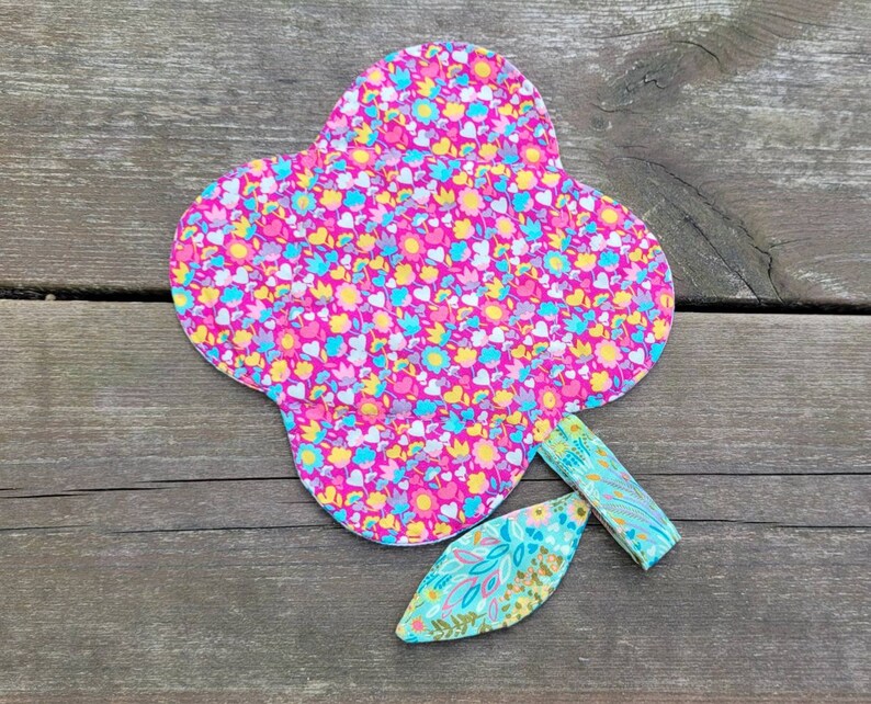 Pu&ograve; includere: Un fiore di stoffa rosa con un motivo floreale e uno stelo di stoffa verde. Il fiore ha la forma di un cuore e un bordo smerlato.