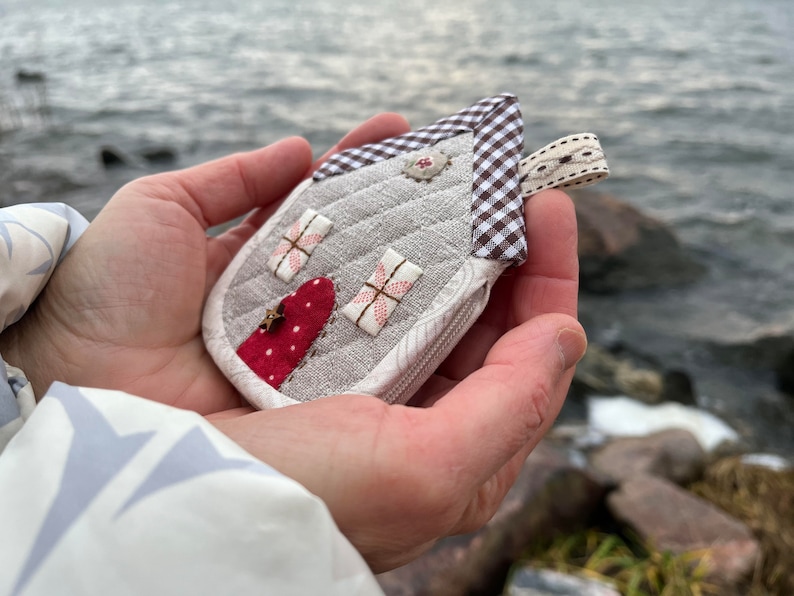 May include: A small, quilted house-shaped zipper pouch with a red heart and floral details. The pouch is made of a light gray fabric with a brown and white checkered trim.