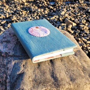 May include: A blue denim journal with a round, embroidered patch featuring a ballerina in pink. The journal is resting on a large, gray rock near a body of water with small rocks along the shore. The journal's pages are visible.