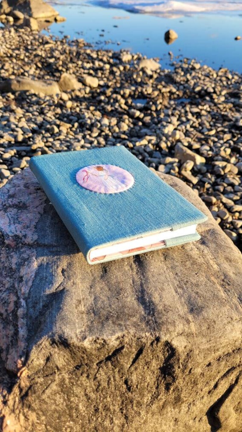 May include: A blue denim journal with a round, embroidered patch featuring a ballerina in pink. The journal is resting on a large, gray rock near a body of water with small rocks along the shore. The journal's pages are visible.