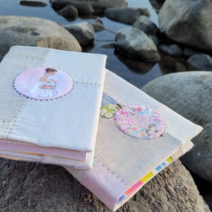 Può includere: Due quaderni fatti a mano con copertine in lino grigio chiaro. Uno presenta un'illustrazione circolare di una ragazza in abito bianco, l'altro un motivo floreale. I quaderni poggiano su una grande roccia vicino all'acqua.