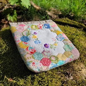 Peut inclure: Une pochette matelassée faite à la main avec un motif floral. La pochette présente un centre blanc avec des fleurs brodées, entouré de patchs hexagonaux colorés et à motifs. La pochette repose sur un lit de mousse verte, avec un arrière-plan naturel flou.