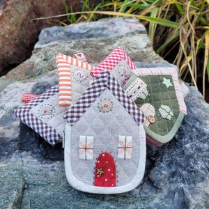 May include: Four small quilted fabric houses in various colors and patterns, including red, white, green, and brown. The houses are arranged on a gray rock surface.
