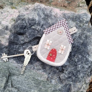 May include: A small, gray house-shaped key chain with a red door and a white star. The house has a gingham roof and is attached to a silver key.