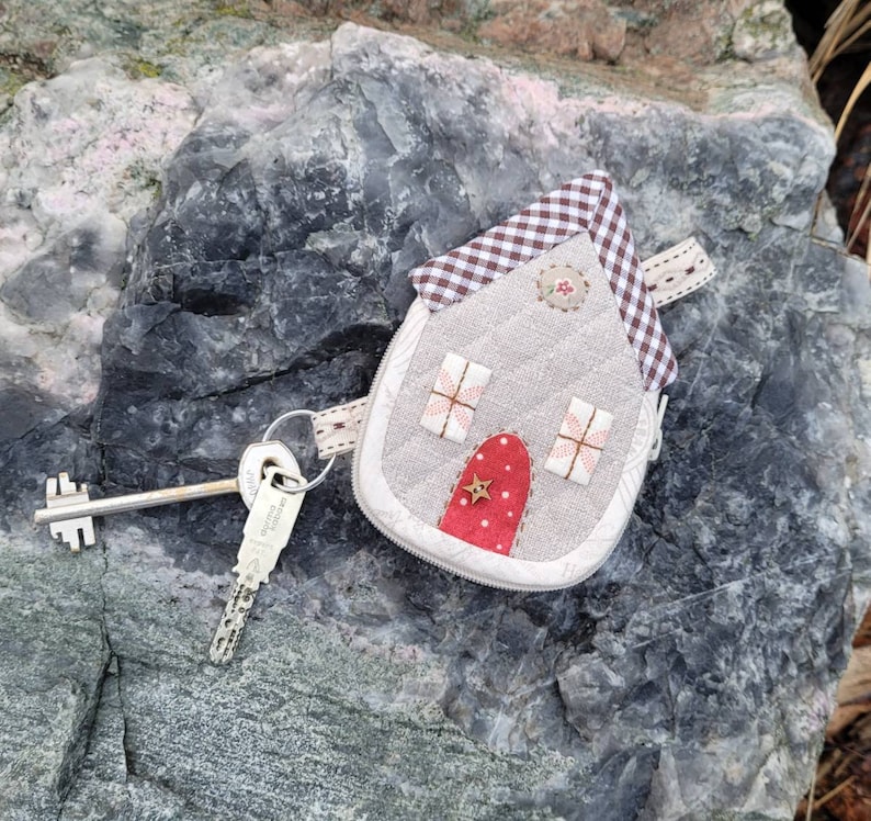 May include: A small, gray house-shaped key chain with a red door and a white star. The house has a gingham roof and is attached to a silver key.