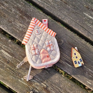 May include: A small, quilted house-shaped keychain with a red heart and the number "2" on the front. The keychain is made of beige fabric with orange and white stripes. A small, yellow house-shaped charm is also included.
