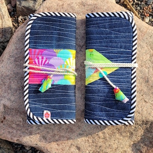 Może przedstawiać: Dwa granatowe etui z tkaniny z białymi przeszyciami i czarno-białą obwódką w paski. Każde etui jest owinięte białym sznurkiem i posiada kolorowy akcent z tkaniny. Etui spoczywają na dużej skale.