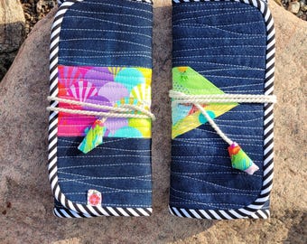May include: Two navy blue fabric cases with white stitching and a black and white striped border. Each case is wrapped with a white cord and features a colorful fabric accent. The cases are resting on a large rock.