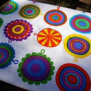 Puede incluir: Tela blanca con un patrón repetitivo de círculos concéntricos de colores. Los círculos son en tonos de rojo, naranja, amarillo, verde, azul y morado. La tela está diseñada por Maija Louekari y se llama "Lappuliida".