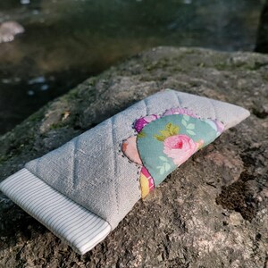 May include: A rectangular quilted fabric case featuring a light gray linen-textured exterior with a white and gray pinstriped end panel. The center is decorated with a layered floral fabric appliqu&eacute; in shades of teal, pink, and yellow.
