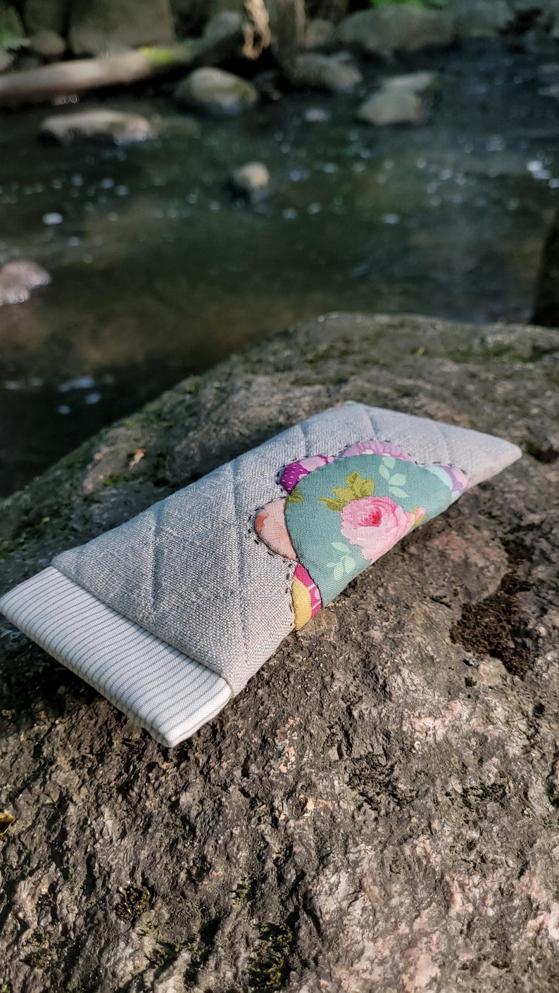 May include: A rectangular quilted fabric case featuring a light gray linen-textured exterior with a white and gray pinstriped end panel. The center is decorated with a layered floral fabric appliqu&eacute; in shades of teal, pink, and yellow.