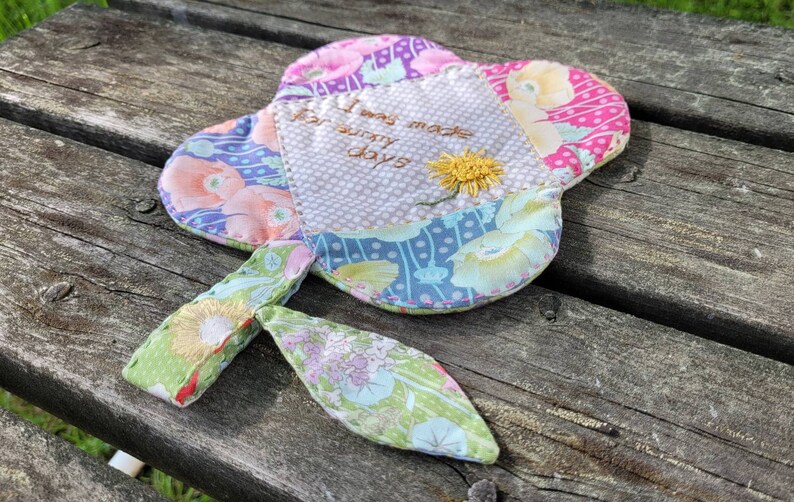 Pu&ograve; includere: Un fiore di stoffa colorato con uno stelo, ricamato con il testo "Sono stato fatto per le giornate di sole".