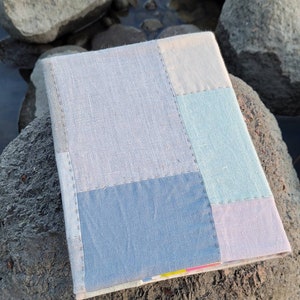 May include: A handmade journal with a patchwork cover. The cover is composed of various pastel-colored linen fabric squares, including light blue, pale green, and lavender. The journal is resting on a large, gray rock near water.