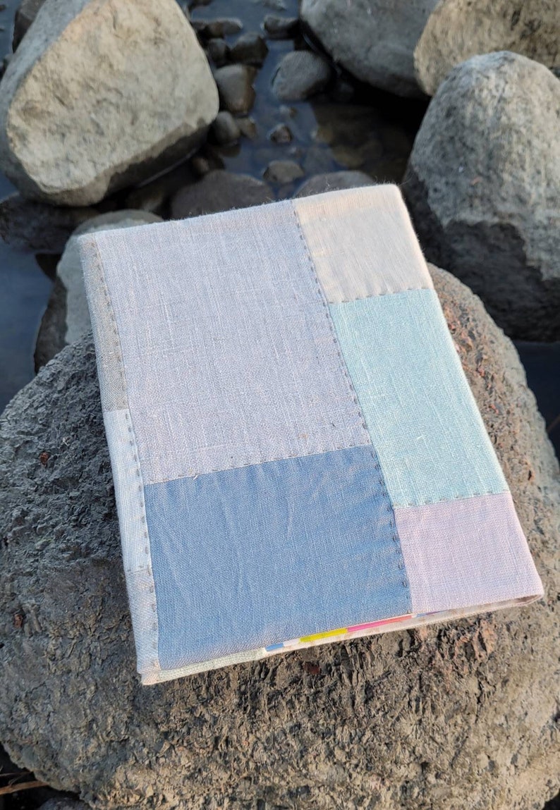 May include: A handmade journal with a patchwork cover. The cover is composed of various pastel-colored linen fabric squares, including light blue, pale green, and lavender. The journal is resting on a large, gray rock near water.