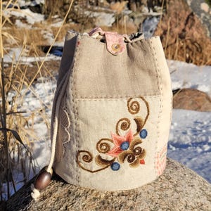 Puede incluir: Una bolsa beige con cordón y diseño floral bordado. La bolsa presenta un parche con un bordado floral en tonos marrón, rosa y azul. La bolsa está hecha de lino y tiene un cierre de botón. La bolsa está sobre una roca.