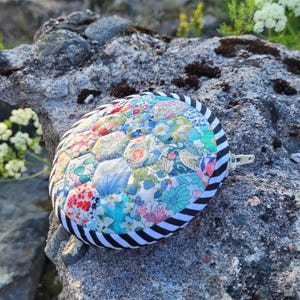 May include: A round, quilted fabric pouch featuring a patchwork of various floral and botanical patterns in shades of blue, pink, green, and yellow. The pouch has a black and white diagonal striped border and a white zip closure on the side.