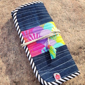 Könnte beinhalten: Eine gerollte Stoffhülle mit einem jeansblauen Hintergrund und einem farbenfrohen Blumen-Patchwork. Die Hülle wird mit einer natürlichen Kordel befestigt und hat einen schwarz-weiß gestreiften Rand. Ein kleiner, dekorativer Knopf ist auf die Ecke genäht.