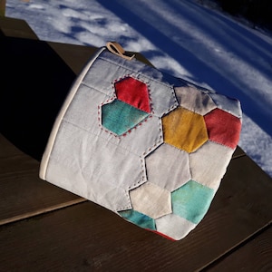 Könnte beinhalten: Eine hellgraue, gesteppte Stofftasche mit einem Patchwork-Design aus bunten, sechseckigen Formen. Die Sechsecke sind in den Farbtönen Rot, Türkis, Gelb und Creme gehalten. Die Tasche hat eine Lederschlaufe und rote Nähte.