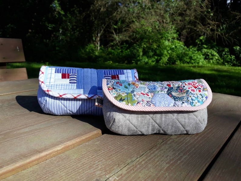 May include: Two quilted fabric pouches. The pouch on the left is blue and white with red accents. The pouch on the right is gray with a floral pattern and a pink and white striped trim.