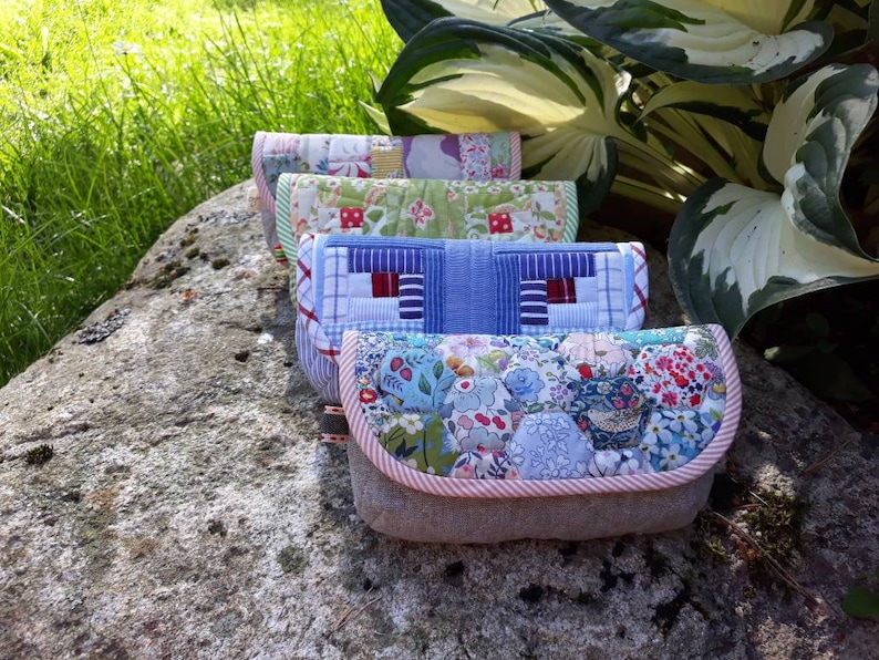May include: Four quilted fabric pouches with different patterns. The pouches are stacked on top of each other on a gray rock. The pouches are made of patchwork fabric with a variety of colors and patterns.