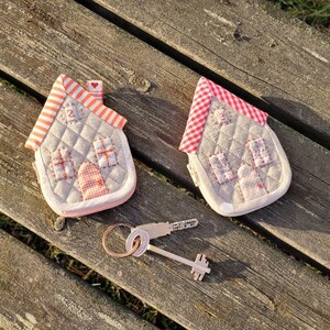 May include: Two small quilted house-shaped keychains with red and white gingham roofs. The keychains are on a wooden surface with a silver key in the foreground.
