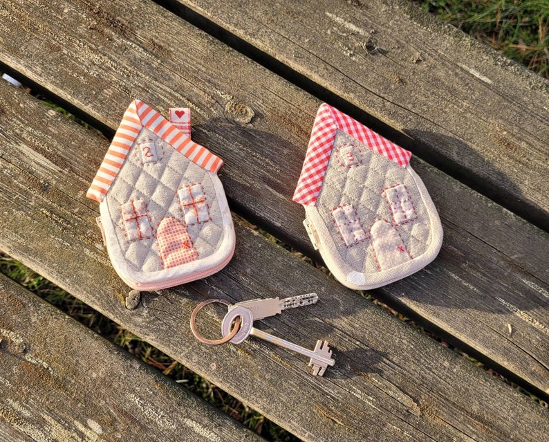 May include: Two small quilted house-shaped keychains with red and white gingham roofs. The keychains are on a wooden surface with a silver key in the foreground.