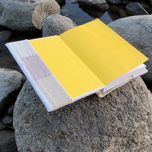 May include: An open book with bright yellow pages rests on a large gray rock near water. The book's cover is a light beige with a lavender and white accent. The pages are a vibrant yellow, creating a striking contrast.