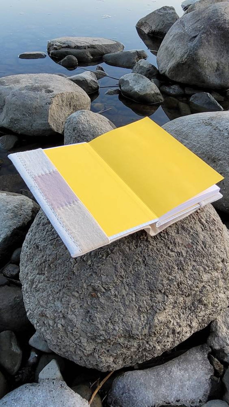 May include: An open book with bright yellow pages rests on a large gray rock near water. The book's cover is a light beige with a lavender and white accent. The pages are a vibrant yellow, creating a striking contrast.