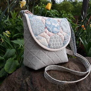 May include: A gray quilted crossbody bag with a patchwork flap featuring a geometric pattern in shades of blue, pink, and white. The bag has a long adjustable strap.