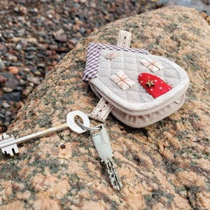 May include: A small, gray, quilted house-shaped keychain with a red door and a gold star. The keychain is attached to a silver key and a silver key ring.