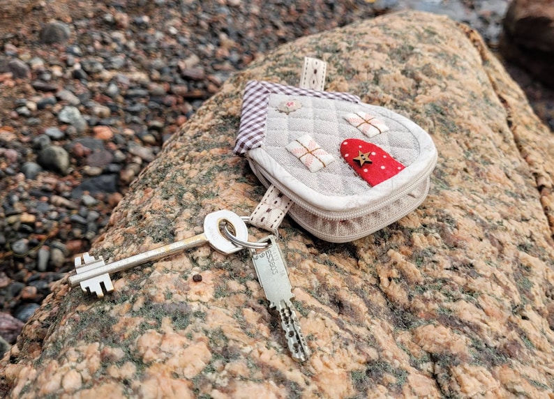 May include: A small, gray, quilted house-shaped keychain with a red door and a gold star. The keychain is attached to a silver key and a silver key ring.