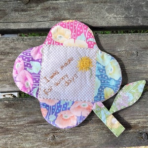 Pu&ograve; includere: Un fiore di stoffa colorato con un messaggio ricamato che dice "Sono stato fatto per le giornate di sole". Il fiore &egrave; fatto di diversi tessuti a motivi e ha uno stelo di foglia verde.