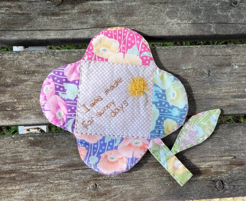 Pu&ograve; includere: Un fiore di stoffa colorato con un messaggio ricamato che dice "Sono stato fatto per le giornate di sole". Il fiore &egrave; fatto di diversi tessuti a motivi e ha uno stelo di foglia verde.