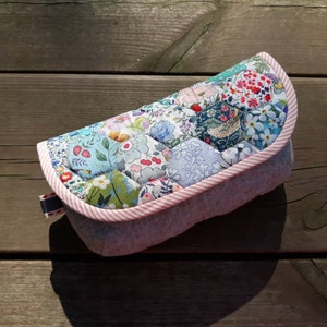 May include: A quilted fabric eyeglass case with a pink and white trim. The case is made of patchwork fabric with a floral pattern.