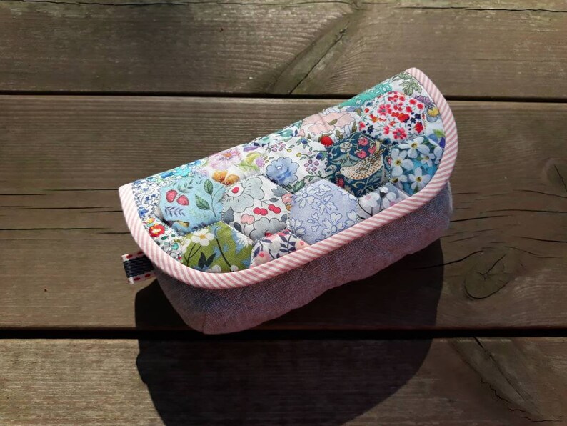 May include: A quilted fabric eyeglass case with a pink and white trim. The case is made of patchwork fabric with a floral pattern.