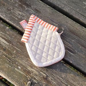 May include: A small, quilted, house-shaped pot holder with orange and white stripes and a pink zipper. The pot holder is made of a soft, beige fabric.