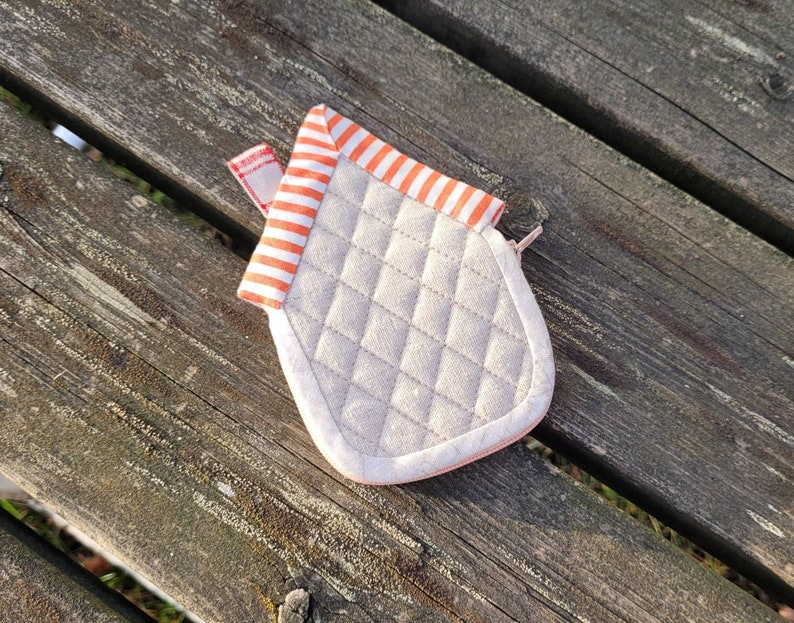 May include: A small, quilted, house-shaped pot holder with orange and white stripes and a pink zipper. The pot holder is made of a soft, beige fabric.