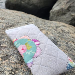 May include: A rectangular quilted fabric case in light gray with a diamond stitch pattern. The front features a decorative scalloped applique made of various floral and polka dot patterned fabric pieces in pink, green, and blue.