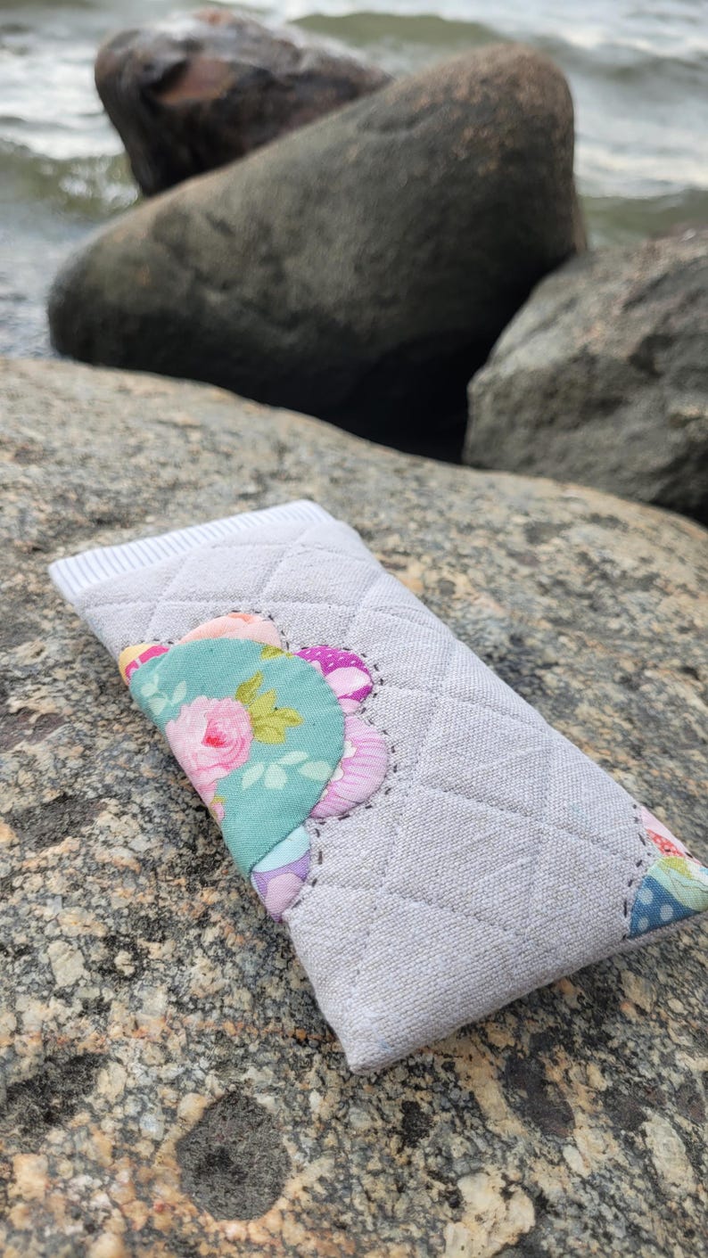 May include: A rectangular quilted fabric case in light gray with a diamond stitch pattern. The front features a decorative scalloped applique made of various floral and polka dot patterned fabric pieces in pink, green, and blue.