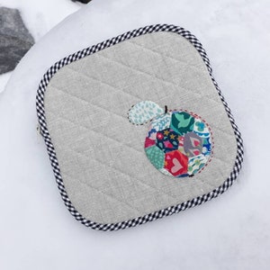 May include: A square, quilted gray fabric pot holder with a black and white checkered border. The pot holder features a colorful, patchwork apple design in the center. The apple has a green leaf and is made of various patterned fabrics.