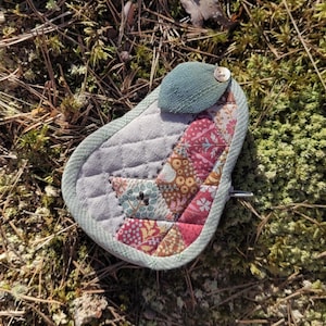 Peut inclure: Un objet matelassé en forme de goutte d'eau avec un motif patchwork de motifs floraux et géométriques rouges, roses et bleus. Il a une base grise matelassée, un rabat en tissu vert et une fermeture à bouton. L'objet repose sur un lit de mousse et d'aiguilles de pin.