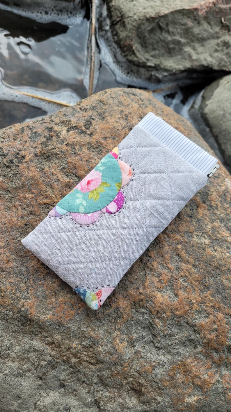 May include: A quilted gray fabric pouch featuring a diamond-stitched pattern and a decorative scalloped edge applique with floral and polka dot prints in shades of pink, blue, and green. The top opening reveals a white and gray striped lining.