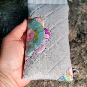 May include: A rectangular quilted fabric pouch in light gray featuring a diagonal stitched pattern. The flap is decorated with layered, scalloped fabric patches in floral and polka dot prints in shades of pink, green, and purple.