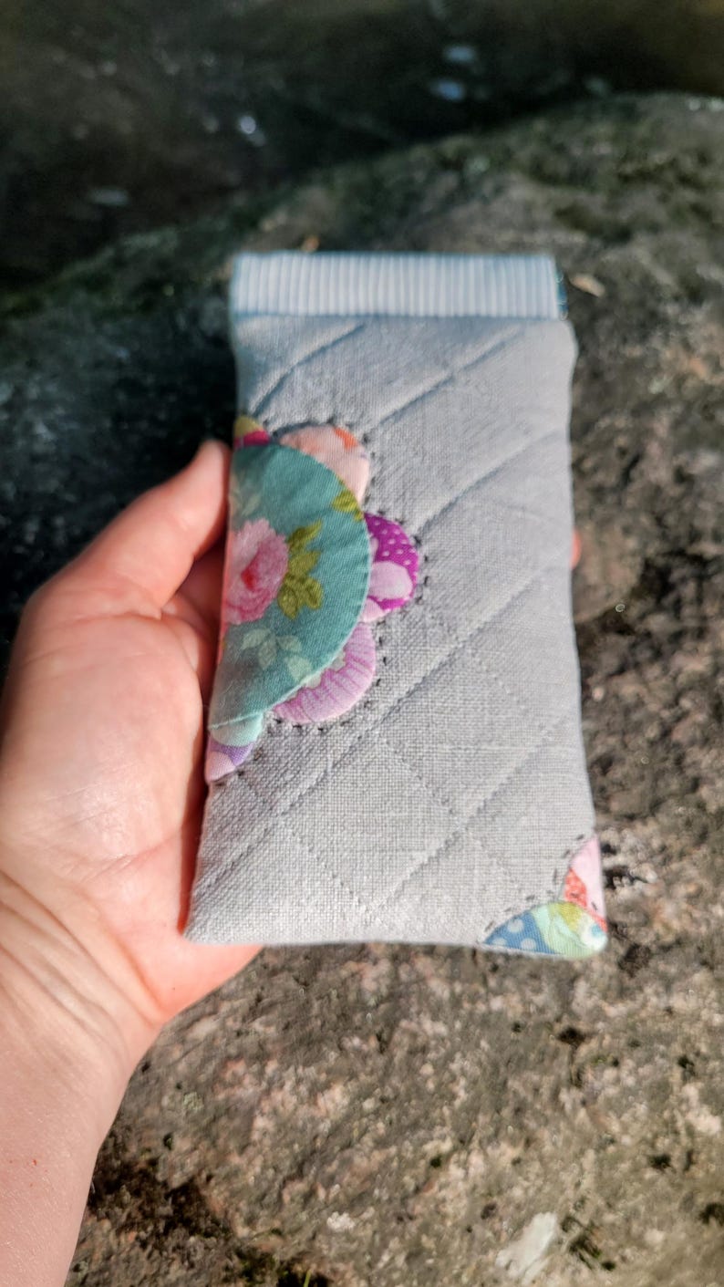 May include: A rectangular quilted fabric pouch in light gray featuring a diagonal stitched pattern. The flap is decorated with layered, scalloped fabric patches in floral and polka dot prints in shades of pink, green, and purple.