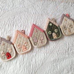 May include: Five small quilted house-shaped pouches in various colors and patterns. The pouches are lined up in a row on a white quilted fabric background.