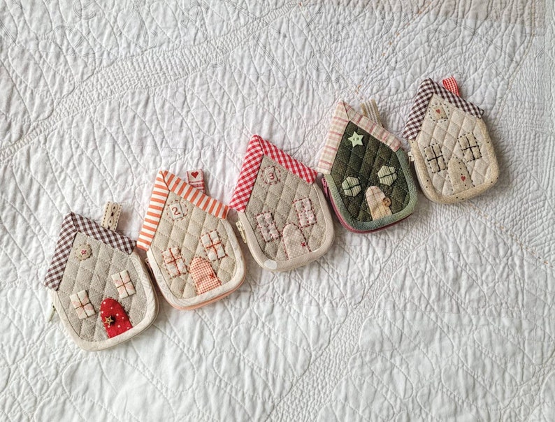 May include: Five small quilted house-shaped pouches in various colors and patterns. The pouches are lined up in a row on a white quilted fabric background.