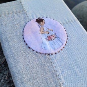 May include: A light blue linen journal with a stitched border. A circular illustration of a woman in a white dress holding flowers is centered on the cover. The background is a soft watercolor wash.