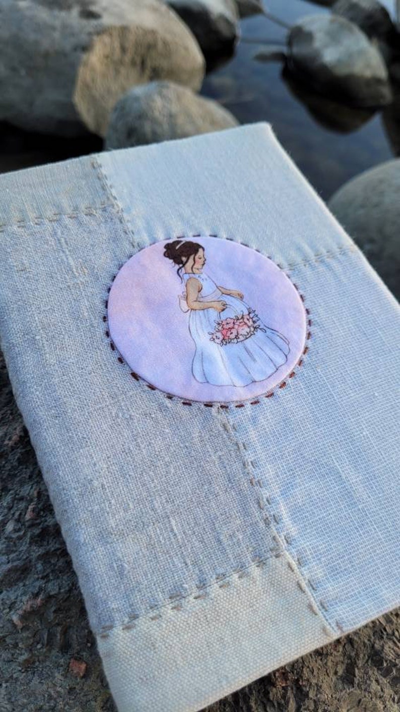 May include: A light blue linen journal with a stitched border. A circular illustration of a woman in a white dress holding flowers is centered on the cover. The background is a soft watercolor wash.