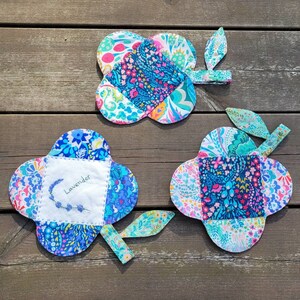 Pu&ograve; includere: Tre sottopentola in tessuto colorati a forma di quadrifoglio con un gambo. I sottopentola sono realizzati in tessuto patchwork con una variet&agrave; di motivi floreali in tonalit&agrave; di blu, rosa, verde e bianco. Un sottopentola ha la parola "Lavender" ricamata.
